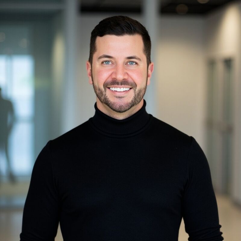 The image shows a man with short dark hair, a neatly trimmed beard, and blue eyes. He is wearing a black turtleneck sweater. He has a warm smile and appears to be in an office setting with a blurred background. The lighting is bright and even, highlighting his features.